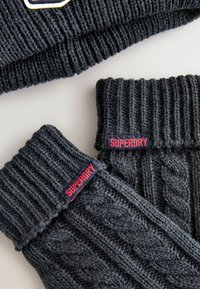 Dunkelgraue Strickmütze und fingerlose Handschuhe mit Rippen- und Zopfmuster. Mit roten "SUPERDRY"-Etiketten an jedem Artikel. Strukturiertes Wollmaterial.