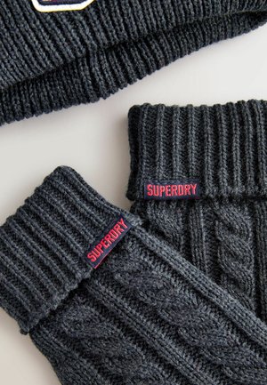 Tumši pelēks pītā adījuma cepure un cimdu pāris bez pirkstiem ar ribotu un kabeli rakstu. Uz katra izstrādājuma ir sarkans "SUPERDRY" etiķešu uzraksts. Tekstūrēts vilnas materiāls.