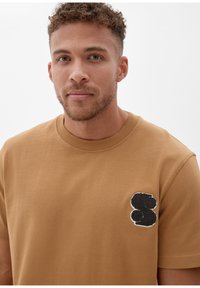 Bruine katoenen T-shirt met korte mouwen, met een gestructureerde zwart-witte logo patch op de linkerkant van de borst. Eenvoudige ronde halsontwerp.