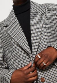 Harmaavalkoinen houndstooth-kuvioitu takki, jossa on jämäkkä kaulus, kaksi suurta ruskeaa nappia ja teksturoitu musta pooloneule alla.