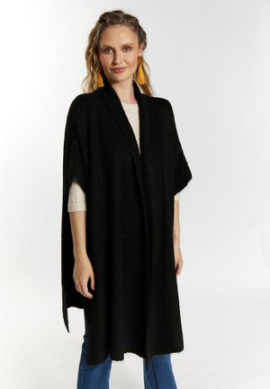 Poncho noir à manches longues en tissu tricoté doux, avec un devant ouvert, une texture côtelée et un ourlet brut. Porté sur un haut de couleur claire et un jean.
