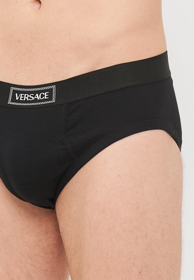 Fekete pamutzokni, bordázott textúrával, széles, "VERSACE" felirattal ellátott elasztikus derékpánttal, kontrasztos fehér varrással.