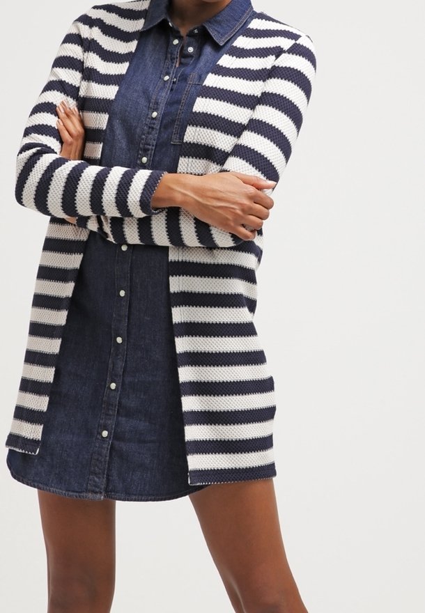 Personne portant une robe en denim boutonnée bleu marine et un cardigan rayé blanc et bleu marine à rayures horizontales, les bras croisés.