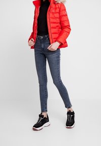 Rød dunjakke med pelsbesat hætte, parret med mørke skinny jeans og sorte sneakers. Jakken har lynlås foran og sidelommer.