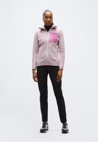 Γυναίκα που στέκεται φορώντας ένα ανοιχτό μωβ υφασμάτινο μπουφάν με φερμουάρ, με ροζ τσέπη στο στήθος, μαύρο παντελόνι και γκρι παπούτσια πεζοπορίας, σε λευκό φόντο.