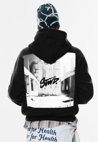 Czarna bluza z kapturem z dużą, białą grafiką przedstawiającą betonową strukturę z czarnym graffiti. Towarzyszy jej wzorzysty beanie.