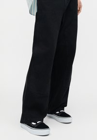 Άτομο που φοράει φαρδιά μαύρη παντελόνια cargo και μαύρα-λευκά αθλητικά παπούτσια χαμηλού τύπου, στέκεται σε λευκό φόντο.