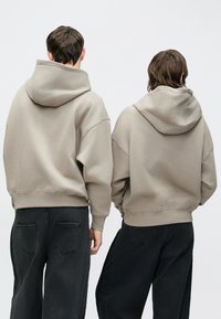 Két személy túlméretezett, texturált szürke kapucnis pulóvereket visel, sötét farmerral kombinálva. A kapucnik laposan a hátukon fekszenek.