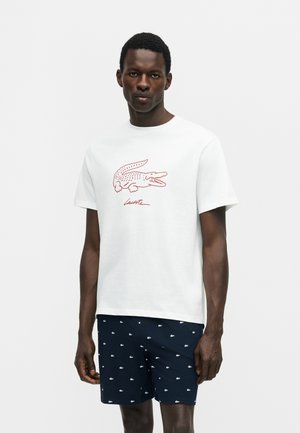 Uomo che indossa una maglietta bianca con un grande logo rosso a coccodrillo e pantaloncini blu navy con piccoli loghi bianchi a forma di coccodrillo, in piedi davanti a uno sfondo semplice.