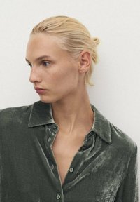 Camisa de terciopelo verde oliva con cuello, cierre de botones en la parte frontal y una textura suave y lisa. Presenta costuras visibles y un corte holgado.