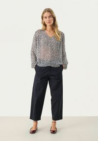 Transparente, gemusterte Bluse mit V-Ausschnitt, kombiniert mit weit geschnittenen, marineblauen Hosen. Die Schuhe sind glänzende bordeauxfarbene Ballerinas mit Knöchelriemen.