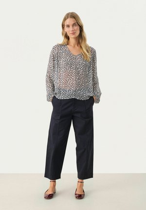 Blusa trasparente e stampata con scollatura a V, abbinata a pantaloni ampî blu navy. Le scarpe sono ballerine lucide bordeaux con cinturini alla caviglia.