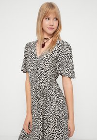 Νεαρή γυναίκα με ίσια ξανθά μαλλιά, φορώντας ένα μαύρο και λευκό φόρεμα με πουά και ένα μαύρο choker με κόκκινο κρεμαστό, κοιτάζοντας πλάγια.