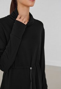 Haut noir à manches longues avec un col drapé, présentant un cordon de serrage à la taille et un accent en métal. Texture lisse et coupe décontractée.