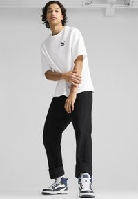 Camiseta blanca de manga corta con un pequeño logo negro, combinada con pantalones negros de puño enrollado y zapatillas gris-azul con acentos blancos.