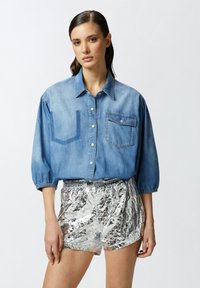 Camicia di jeans blu chiaro con maniche lunghe e a sbuffo, due tasche sul petto e pantaloni corti argentati stropicciati. Chiusura con bottoni e texture morbida.