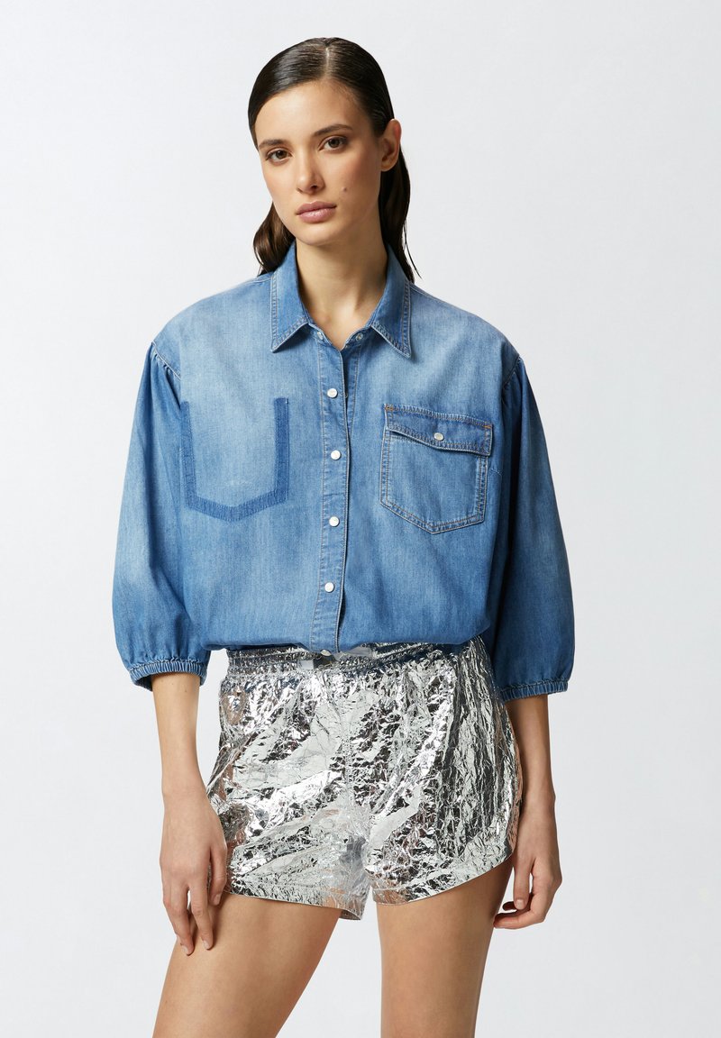 Camicia di jeans blu chiaro con maniche lunghe e a sbuffo, due tasche sul petto e pantaloni corti argentati stropicciati. Chiusura con bottoni e texture morbida.