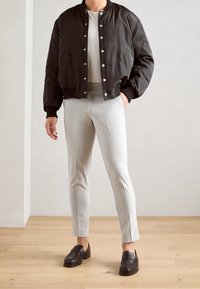 Svart bomberjacka med silverknappar, vit t-shirt, ljusgrå skräddarsydda byxor och svarta loafers på ett trägolv mot en neutral bakgrund.