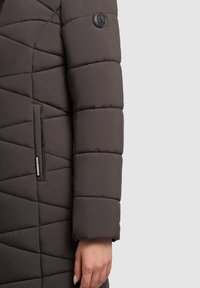 Donkergrijze gewatteerde jas met een chevron-quiltontwerp, zijzak met rits en een ronde logo patch op de linkermouw. Gladde textuur.