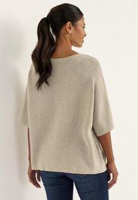 Frau mit dunklem Pferdeschwanz, die ein beiges kurzärmeliges Strickoberteil und blaue Jeans trägt, seitlich vor einem schlichten hellen Hintergrund stehend.