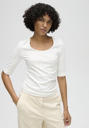 Donna con capelli ricci che indossa una maglietta bianca a mezza manica e pantaloni beige, in piedi con una mano in tasca davanti a uno sfondo semplice.