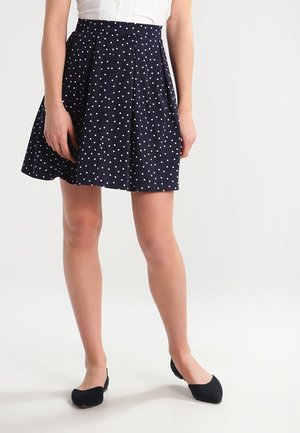 Femme portant une jupe plissée bleu marine à pois blancs, associée à un haut blanc et des ballerines noires, se tenant sur un fond clair.