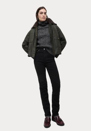 Giacca cerata verde con colletto marrone, maglione grigio a trecce, jeans neri e mocassini bordeaux. Vestibilità su misura e dettagli testurizzati.