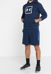 Uomo che indossa un cappuccio navy blue di Under Armour, pantaloni corti sportivi abbinati con un motivo laterale, calze nere e sneakers nere, in piedi con le mani in tasca.