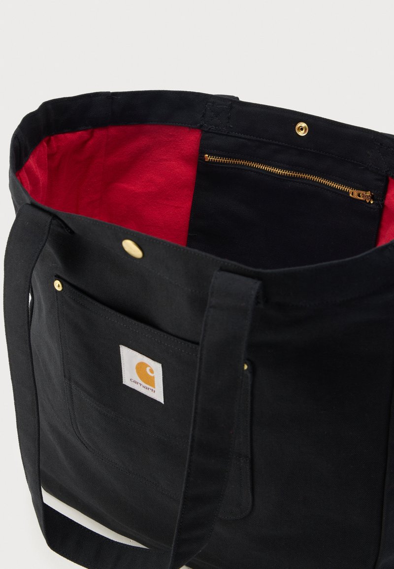 Black canvas tote bag with red inner lining, gold zipper pocket inside, front pocket with Carhartt logo patch, and gold snap closure.