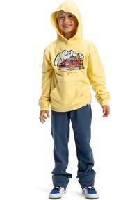 Uśmiechnięte dziecko w żółtej bluzie z kapturem z grafiką czerwonego samochodu, w niebieskich spodniach i białych trampkach z niebieskimi akcentami, stojące na białym tle.