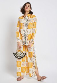 Yellow and white patterned two-piece outfit with oversized shirt and wide-leg pants, featuring text and floral designs. Black beaded handbag.