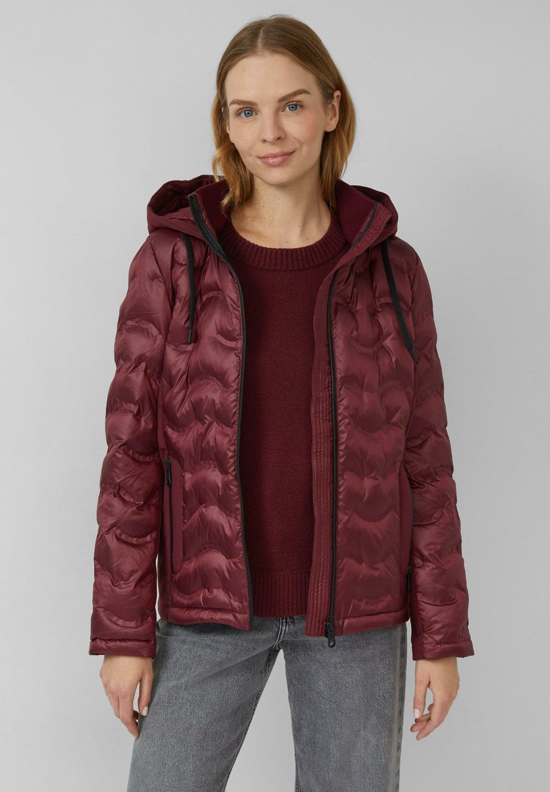 Veste rembourrée bordeaux avec capuche, présentant un design matelassé, des poches latérales et des accents noirs. Portée par-dessus un pull assorti.