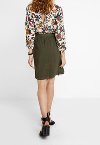 Blouse florale à manches longues, décolleté en V et ouverture au dos ; jupe verte olive ; sandales à talons noires avec brides à la cheville.