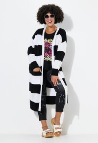 Cardigan largo a rayas en blanco y negro, con puños acanalados, combinado con una camiseta gráfica negra y leggings negros con estampado, complementado con unas gafas de sol.