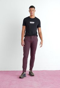 Crna pamučna majica s bijelim "Levi's" logotipom, u kombinaciji s maroon slim-fit hlačama i patikama s kamuflažnim uzorkom na ružičastom tepihu.