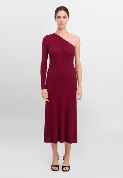 Robe en tricot côtelé bordeaux avec une longue manche et une épaule dénudée, ajustée au corps, s'évasant doucement à l'ourlet, accompagnée de talons noirs.