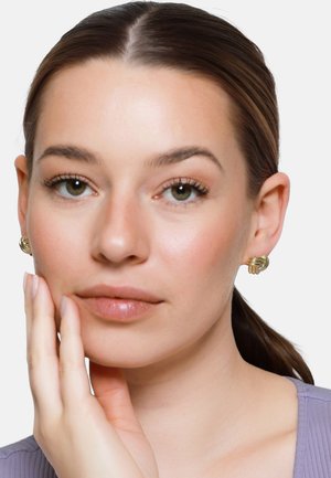 Młoda kobieta o jasnej cerze i brązowych, związanych z tyłu włosach, nosząca złote kolczyki w kształcie supła i lawendową bluzkę, opierająca twarz na dłoni.