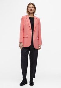 Coral blazer with a single button and two front pockets, paired with striped black trousers and black shoes. Smooth fabric, tailored fit.