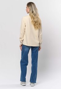 Bēša ar garām piedurknēm topiņš ar apaļu kakla izgriezumu, kopā ar augstā vidukļa ziliem džinsiem un baltām sporta kurpēm, uz neitrāla fona.