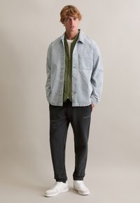 Hellblaue Jeansjacke über einem grünen, zum Zuknöpfen geeigneten Shirt, kombiniert mit schwarzen Jogginghosen und weißen Sneakers. Die Jacke hat eine Vordertasche und Knöpfe.