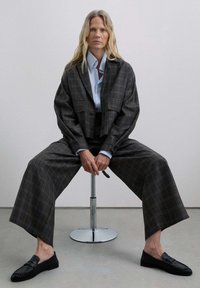 Chaqueta de cuadro grande en gris, camisa de un azul claro y pantalones de cuadro de pierna amplia. Mocasines negros con detalles texturizados. Diseño simple y moderno.
