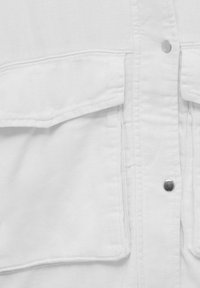 Biała bawełniana kurtka z klapkową kieszenią po lewej stronie i dwoma metalowymi napami wzdłuż przedniego szwu.