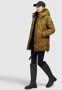 Olive-green quilted puffer jacket with a high collar and zippers, paired with black leggings and knee-high black boots.