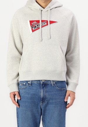 Homme portant un sweat à capuche gris clair avec un logo triangulaire rouge et blanc indiquant "Gant USA" et un jean en denim bleu, debout devant un fond blanc.