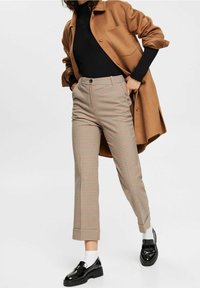 Brązowy, oversize'owy płaszcz, czarny prążkowany sweter, kraciaste spodnie w beżowe, czerwone i szare wzory, o dopasowanym kroju, do tego czarne buty i białe skarpetki.