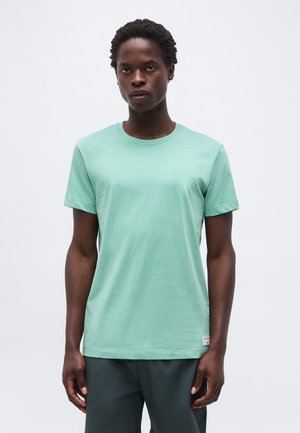 Młoda osoba o krótkich kręconych włosach, ubrana w jednolity, jasnozielony t-shirt z krótkim rękawem i ciemne spodnie na jednolitym tle.