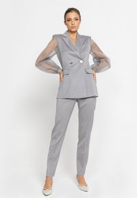 Szary dopasowany garnitur z długimi, przezroczystymi bufiastymi rękawami, podwójnymi guzikami i dopasowanymi spodniami, uzupełniony o beżowe buty w szpic.