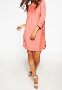 Donna in un vestito corallo rosa a maniche tre quarti, lungo fino al ginocchio, con tacchi aperti bianchi a caviglia, in piedi davanti a uno sfondo bianco.