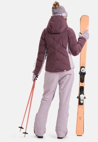 Bordeauxfarbene Pufferjacke mit Kapuze, helllila Schneehosen, rosa Skistöcke und Skier mit orangefarbener Oberseite und schwarzen Bindungen.
