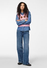 Chaleco de punto rosa con patrones de flores sobre una camisa de denim azul, combinado con unos vaqueros acampanados azules y zapatos negros.